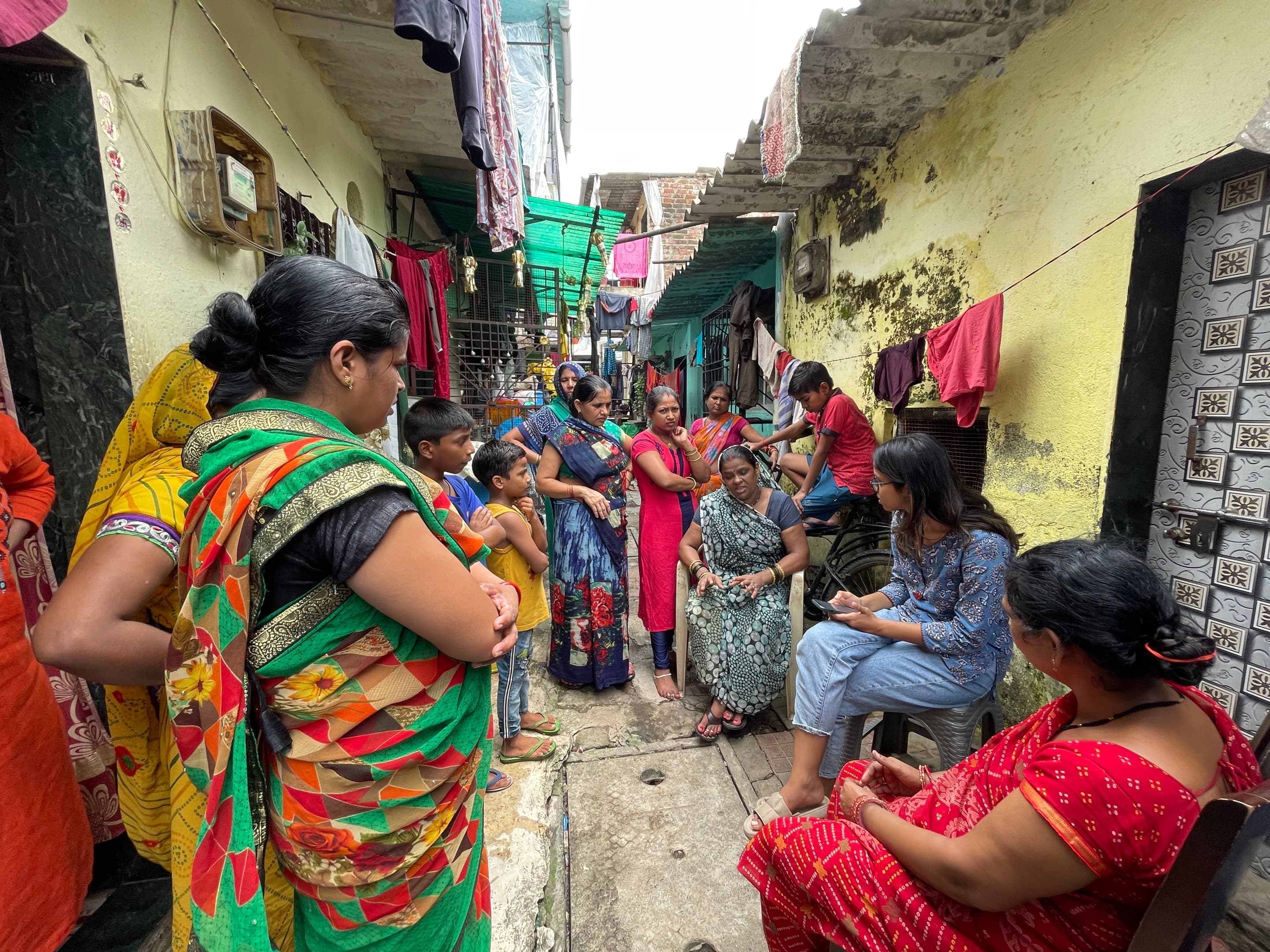 Anushka Shahdadpuri listens at a community engagement session. A group of women wearing brightly colored saris are gathered around her in a narrow alleyway. 