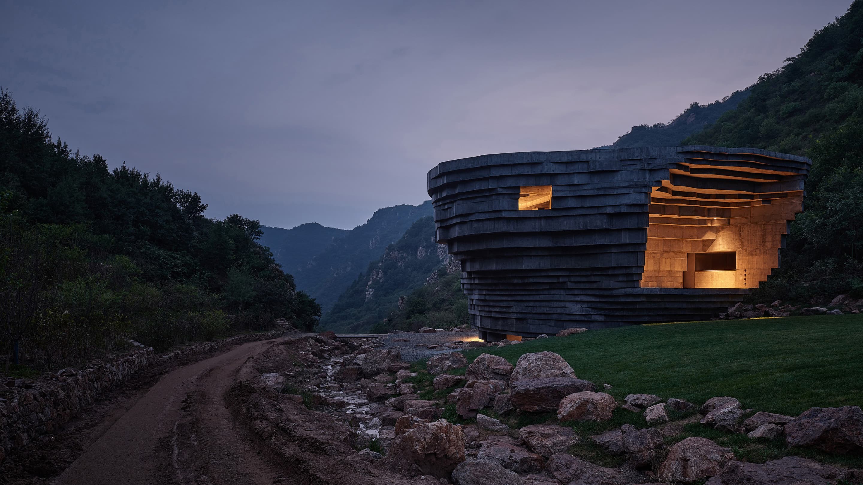 A concrete building with rough-hewn walls lit from within with a golden light against a background of forest-covered mountains.