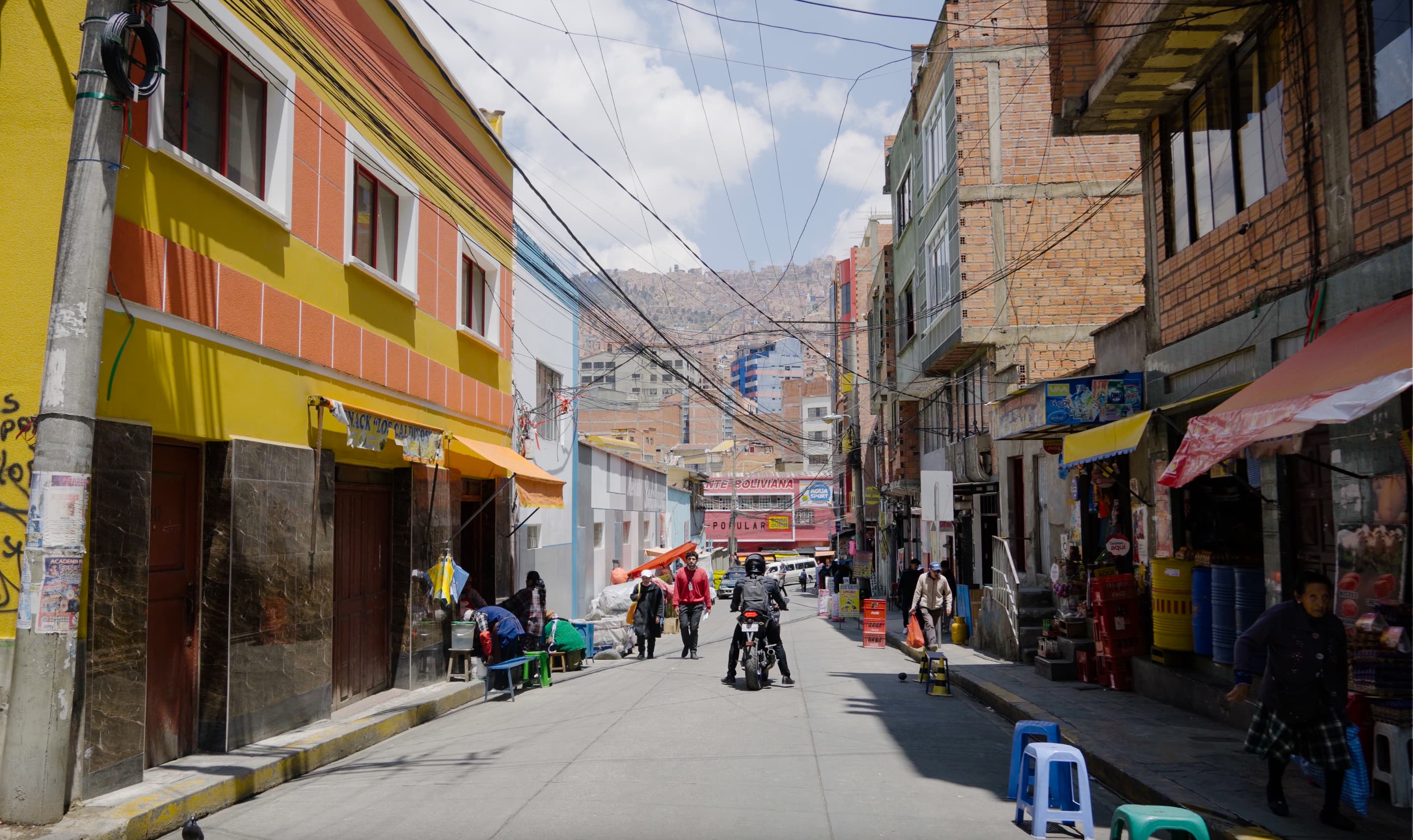 A vibrant street in El Alto. A dense swathe of brick buildings climb up the Bolivian Andes in the background. 