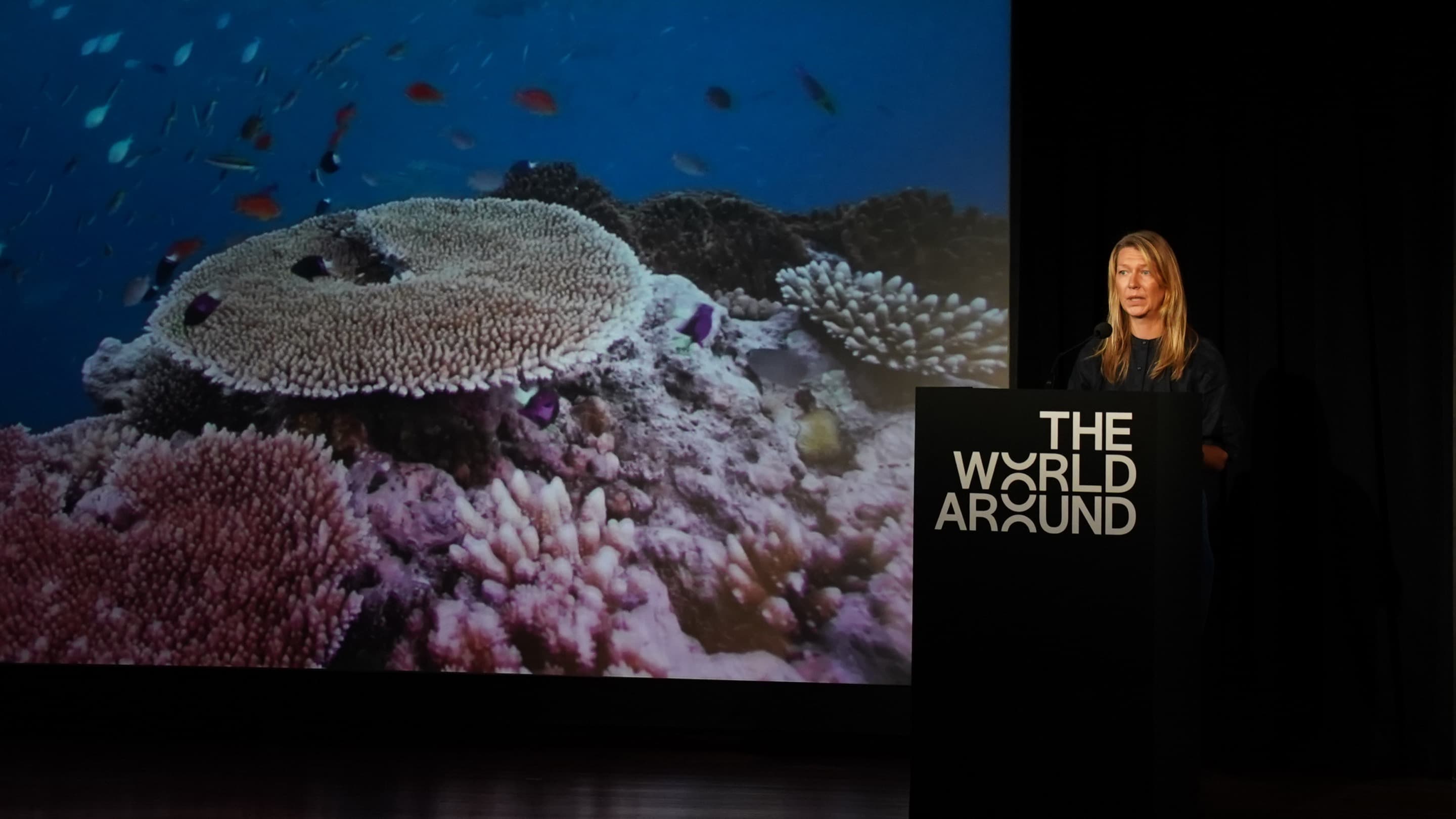Pirjo Haikola speaks in front of a photo of a coral reef at The World Around Summit 2023.