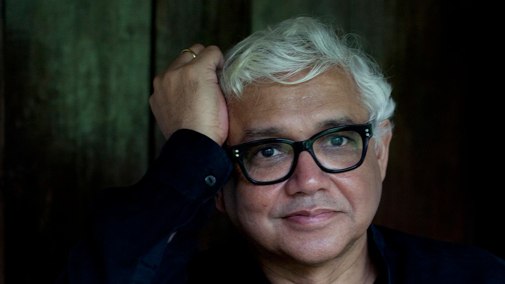 Amitav Ghosh rests his head against his hand in front of a green background.