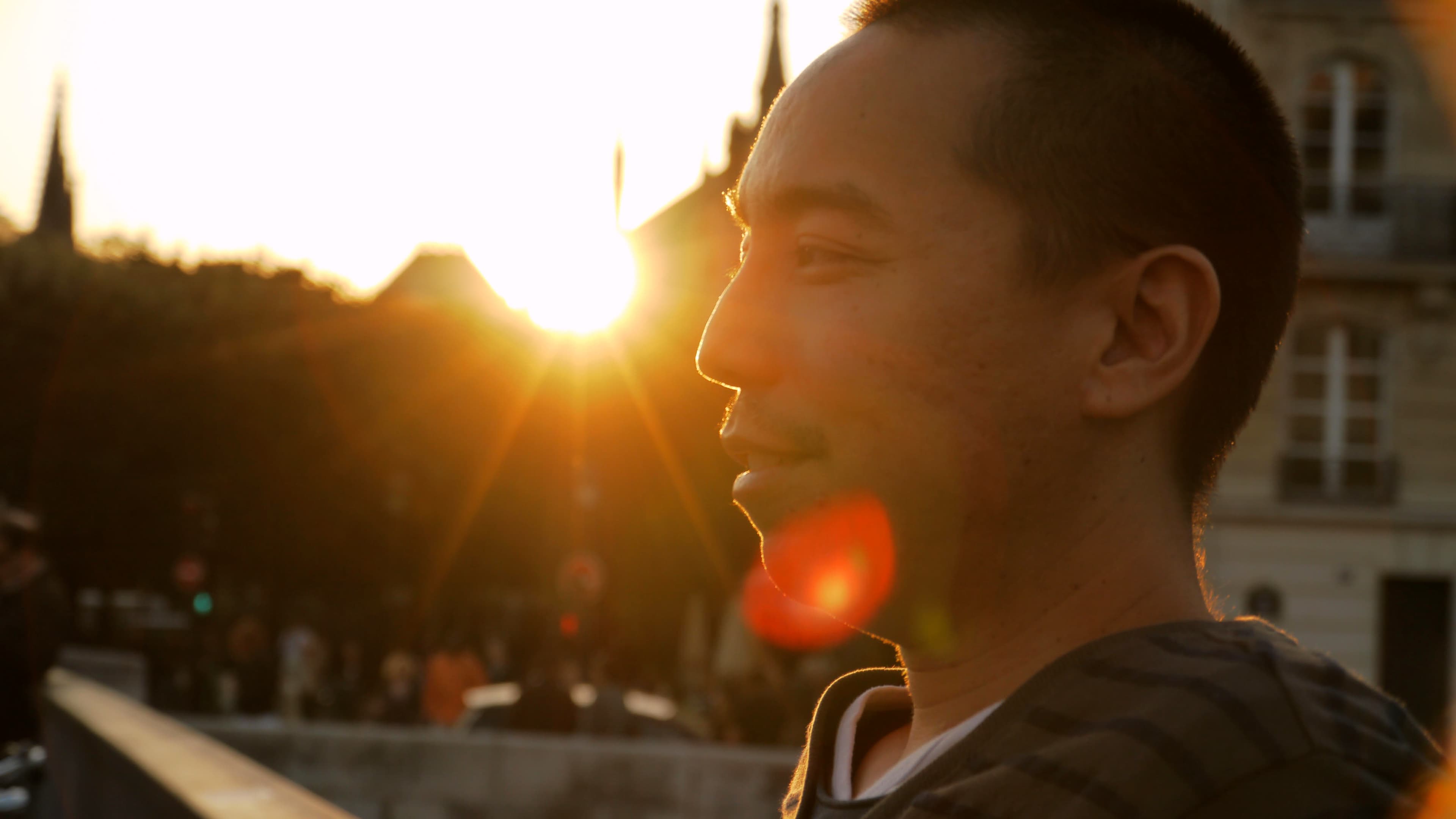 Apichatpong Weerastethakul in profile. The sun sets behind buildings in the background.