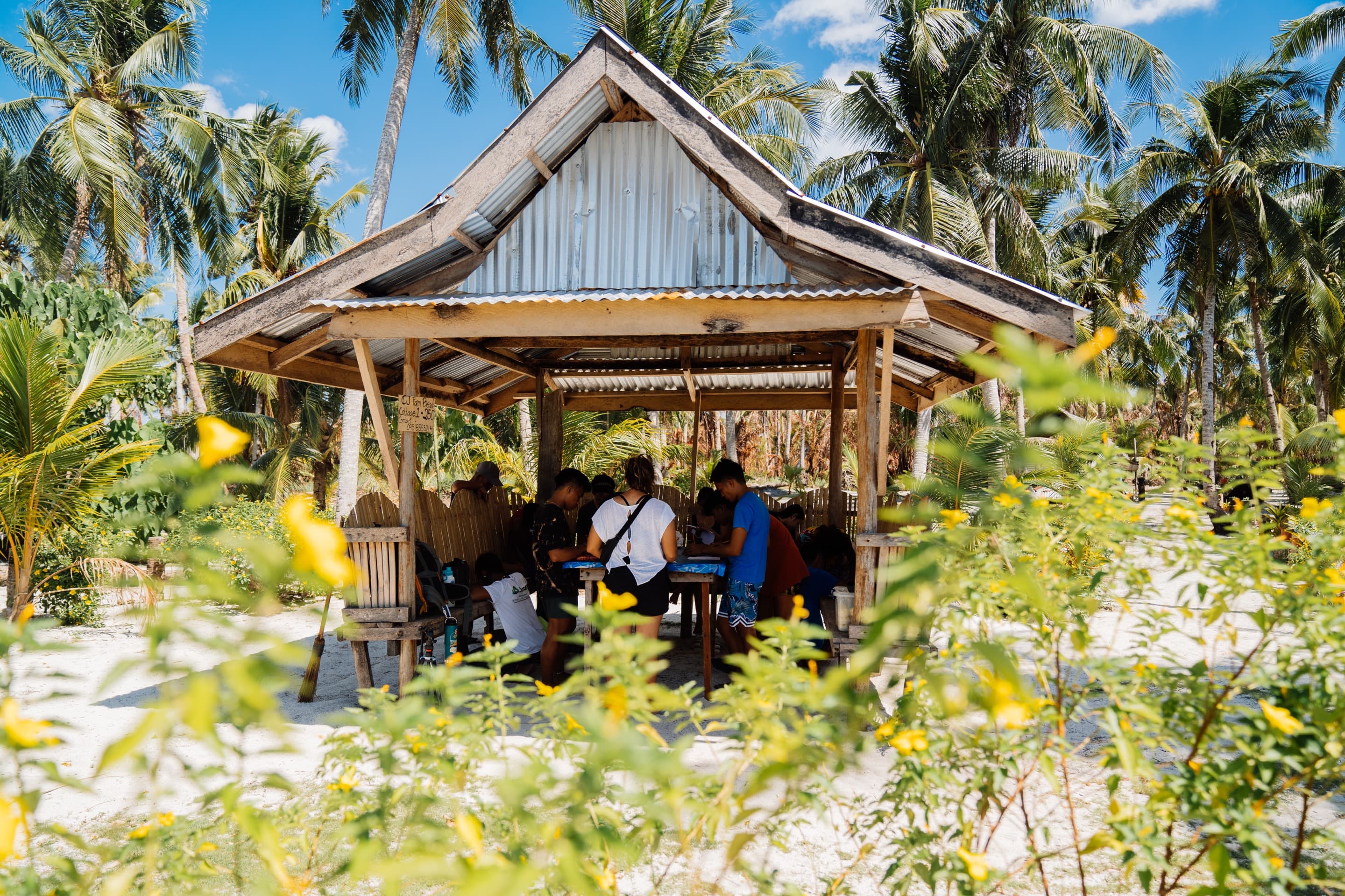 Young people gather around a table in a wooden structure with a corrugated steel roof. Palm trees stand tall in the background.