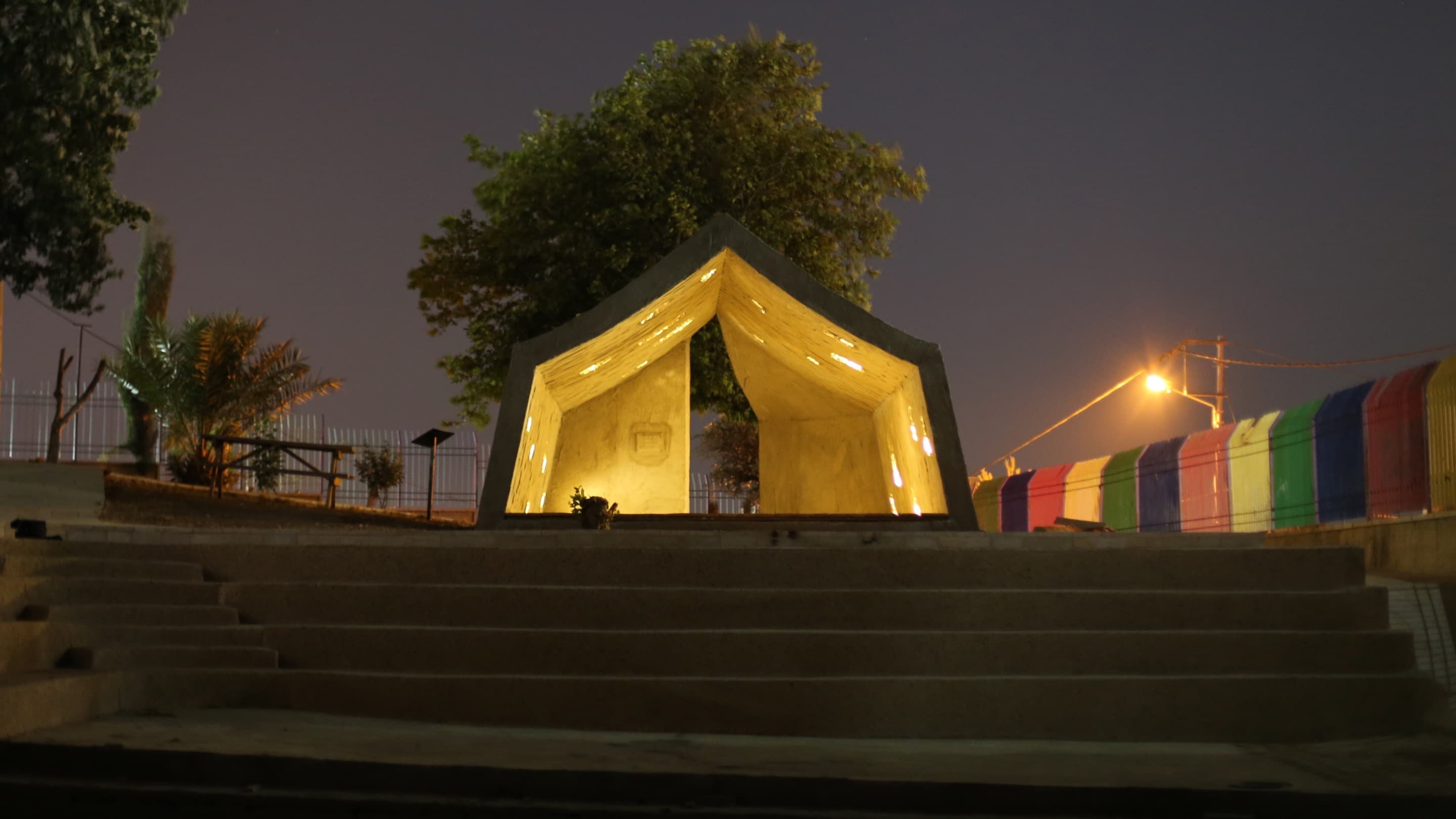 Stairs lead up to a concrete tent lit with a golden light from within. The structure stands against the night sky, with a multicoloured fence and a bright street light in the background.