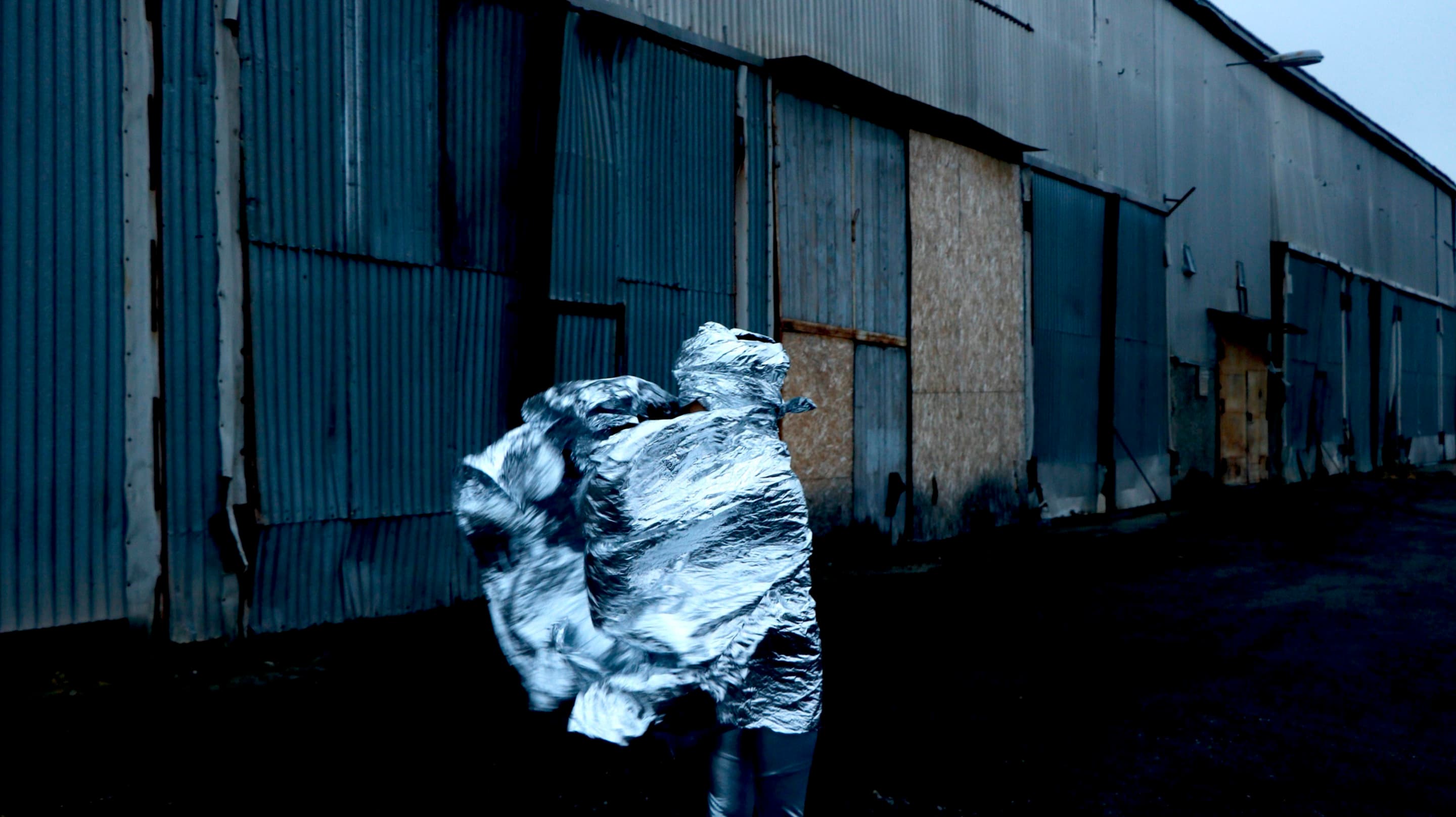 A person cloaked in a reflective heat blanket runs past a ramshackle warehouse in a still from Himali Singh Soin's film We Are Opposite Like That.