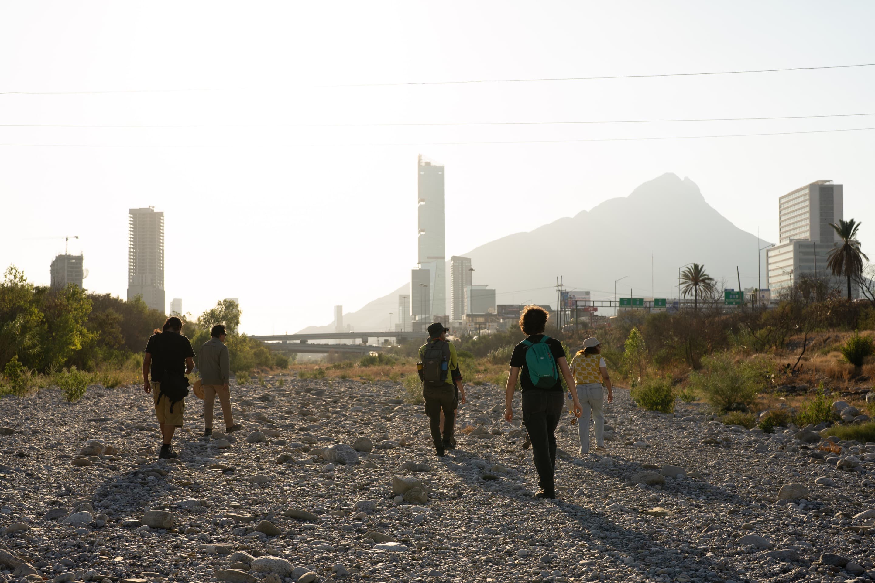 Viaje Al Microcosmos lead a group along the dried-up river bed of the Santa Catarina River in Monterrey, Mexico. Buildings rise in front of mountains in the background.
