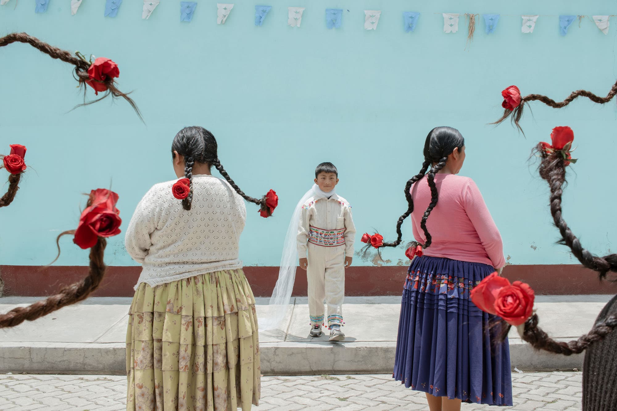 A boy in traditional Bolivian dress and a princes cloak walks towards women with roses at the end of their braids in a still from Warawar Wawa.