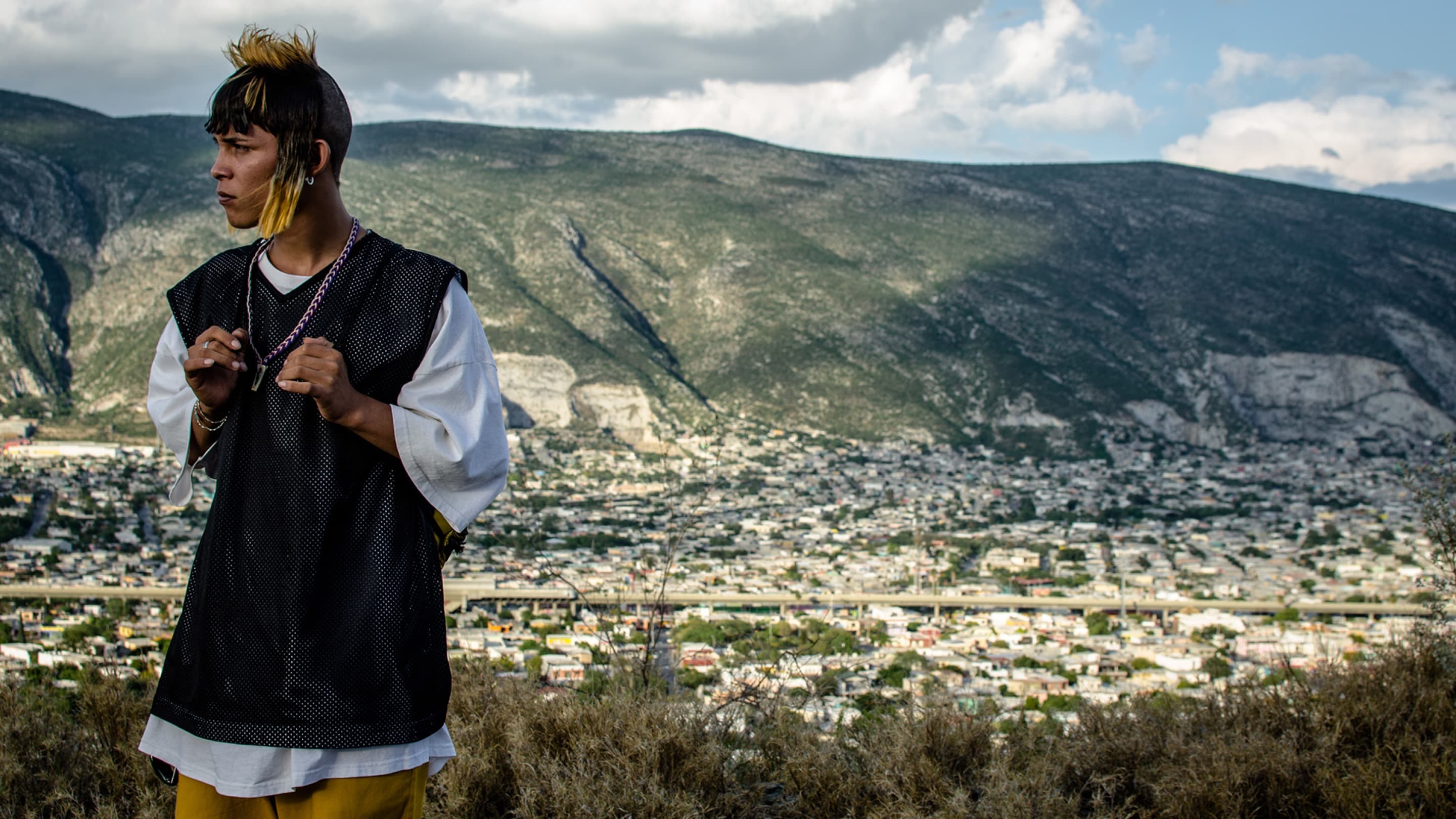 Still from Ya No Estoy Aqui, 2019 by Fernando Frías. A boy wearing a T-shirt and a mesh waistcoat stands looking to the side. Clouds drift over mountains and low-lying residential buildings in the valley below in the background.